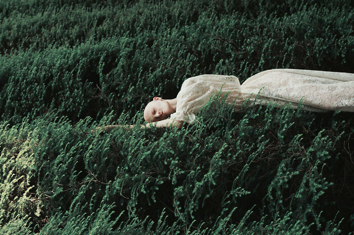 Unique model in a white dress lying on lush green foliage, blending with the natural surroundings.