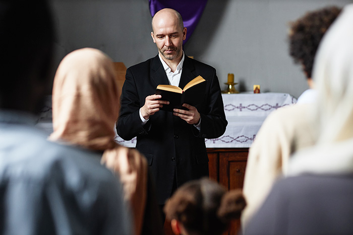 Religious leader reads to congregation, holding a book, standing in front of an altar, people listening attentively.