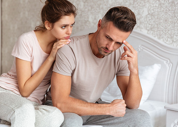 Wife comforts husband on bed, both wearing casual clothes, highlighting an emotional moment.