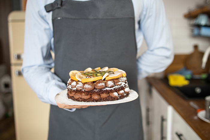 Man holding a chocolate cake with fruit on top, wearing a blue shirt and apron, in a kitchen setting.