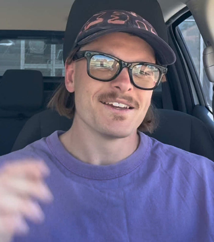 Podcaster in a car wearing glasses and a cap, smiling while discussing a topic related to "Not All Men.