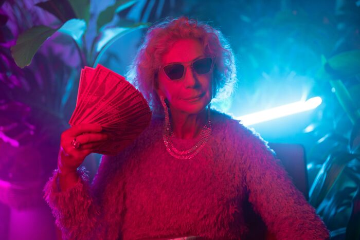 Elderly woman in sunglasses holding money, showcasing her life cheat codes in a vibrant, neon-lit setting.