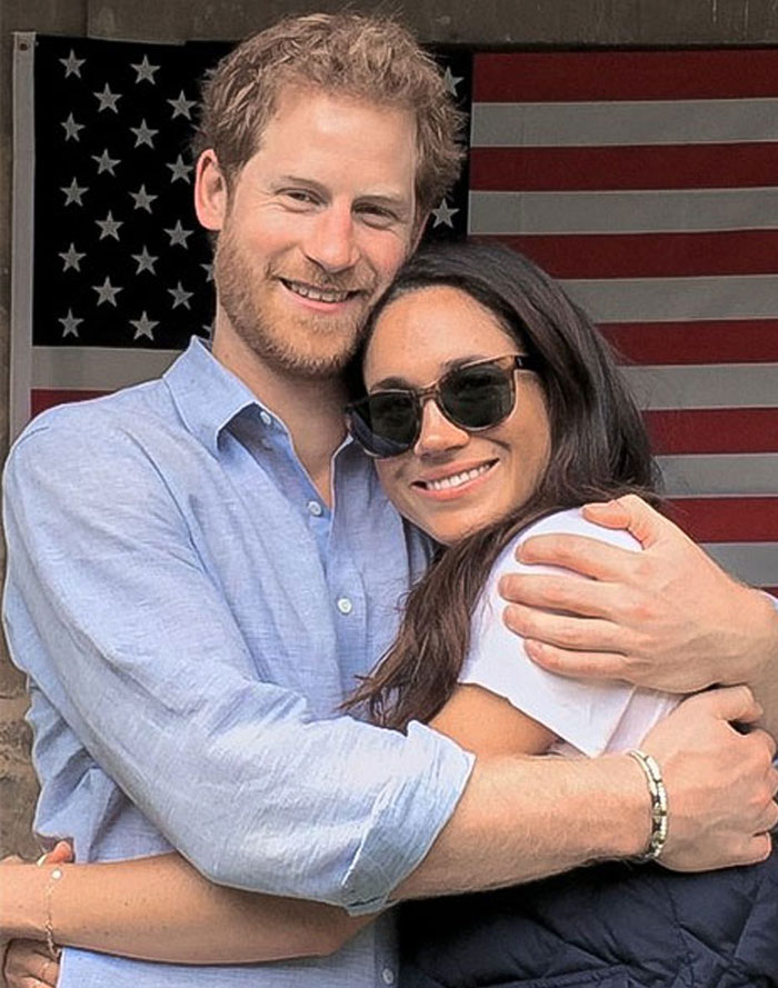 Couple embracing in front of the American flag, Meghan Markle wearing sunglasses.