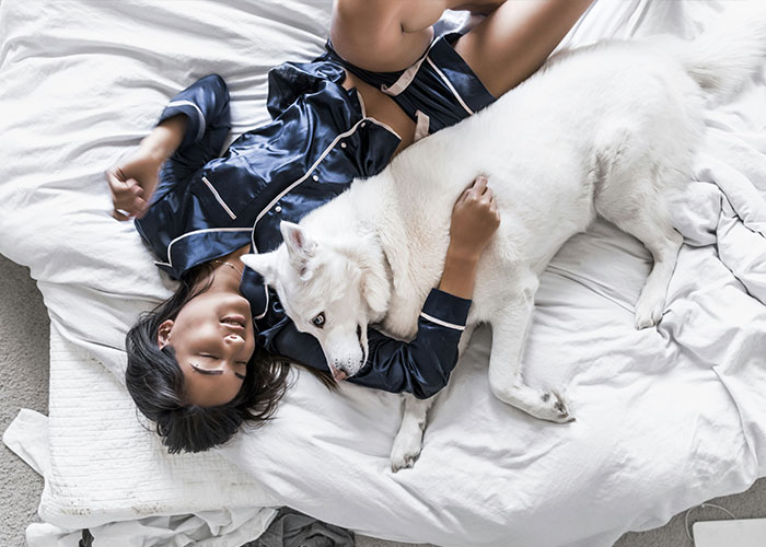 Woman in silk pajamas cuddling a large white dog on a bed, adding a whimsical touch to her day.