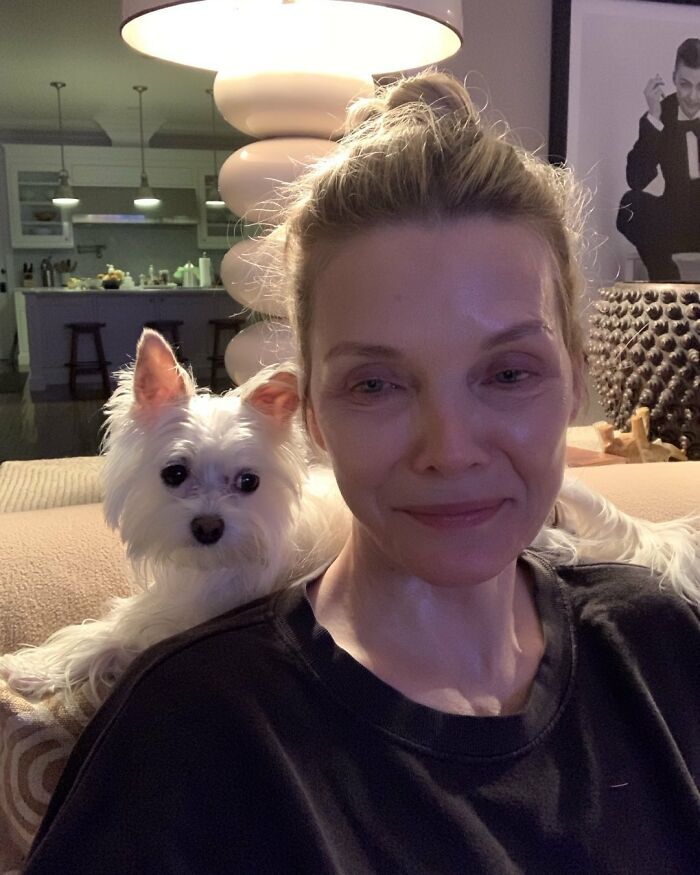 A person relaxing at home without makeup, sitting with a small white dog.