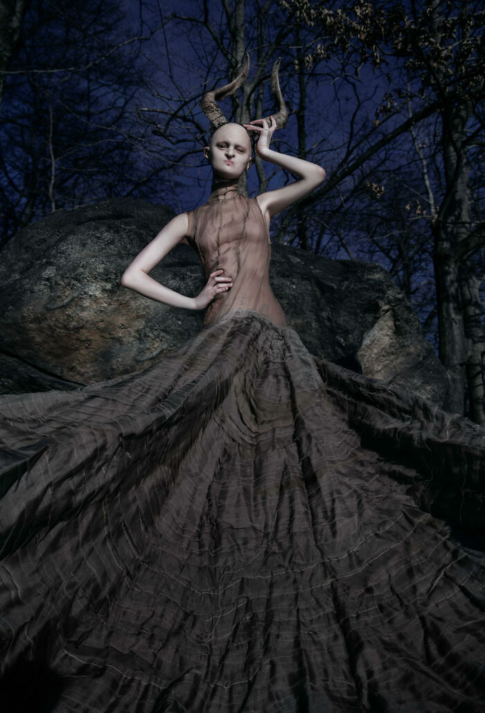Unique model in an extravagant gown with horn accessories, posing dramatically against a forest backdrop at dusk.