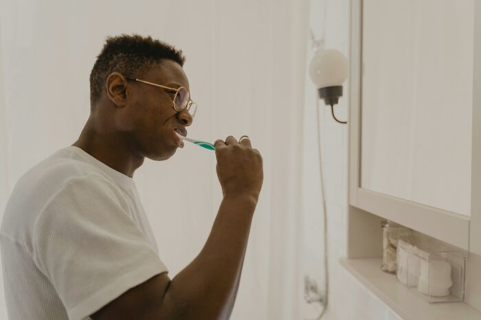 Person brushing teeth in a bathroom, wearing glasses and a white shirt, highlighting deep weird flags in routines.