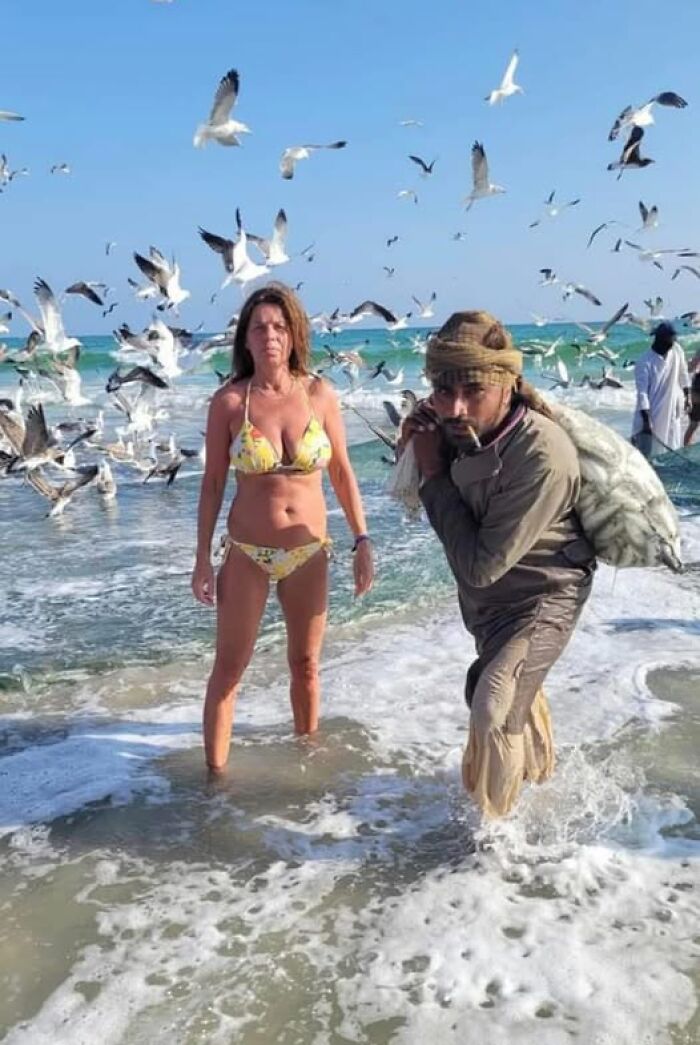 Weird image of a woman in a bikini and a man with a sack surrounded by seagulls at the beach.