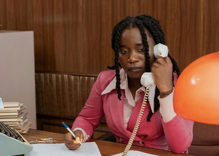Person in a pink suit on the phone, sitting at a desk with papers, symbolizing revenge against bosses.