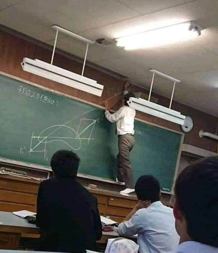 Teacher standing on a table, drawing on a high chalkboard while students watch, creating a weird image in a classroom setting.