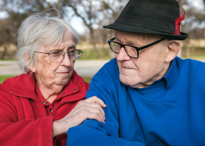 Elderly couple seated outdoors, sharing a heartfelt moment, the woman comforting the man by touching his arm.