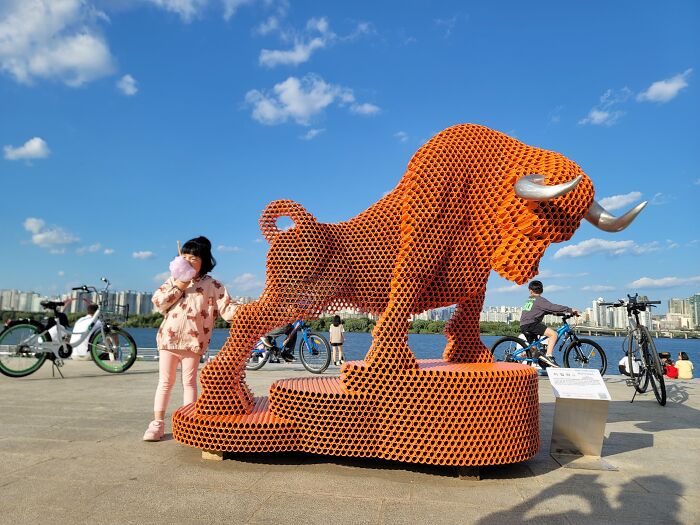 A child stands by a metal pipe sculpture of a bull by Yi Chul Hee, set against a cityscape and blue sky.