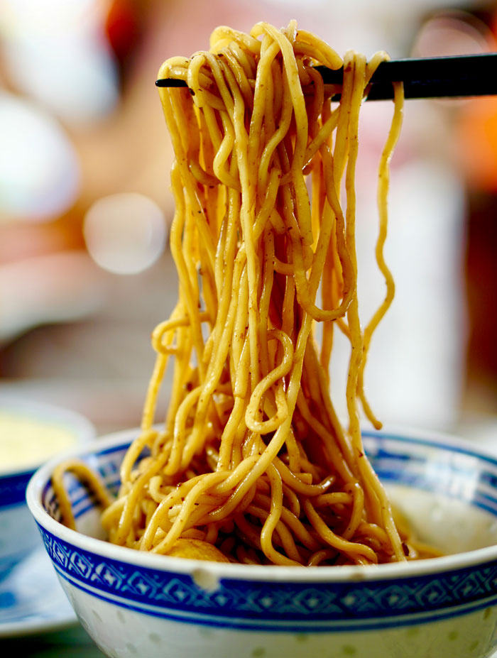 Chopsticks lifting noodles from a decorative bowl; concept of how food can be viewed as trashy if poor, classy if rich.