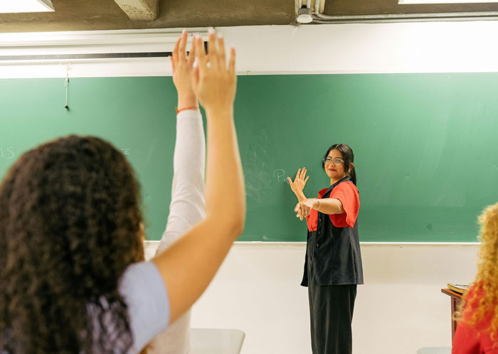 Profesora sonriendo frente a estudiantes levantando la mano en un aula con pizarra.