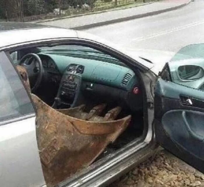 Weird image of a car with a large excavator bucket inside, appearing through the passenger door on a city street.