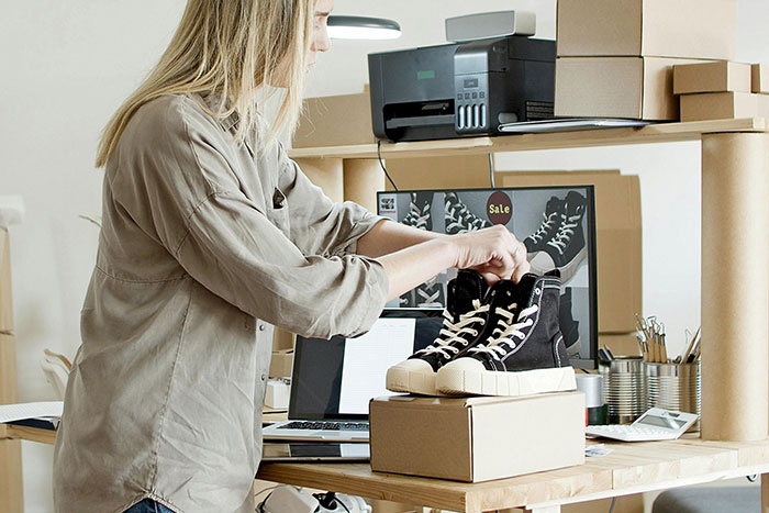 Woman packing sneakers in a home office, exploring safe side hustle ideas with boxes and a laptop nearby.