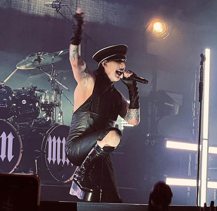 Singer performing energetically on stage in dramatic attire, with drums and bright stage lights in the background.