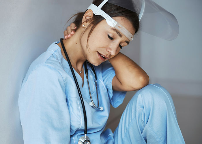 Nurse in blue scrubs and face shield, sitting tiredly against a wall, reflecting on heartfelt patient experiences.