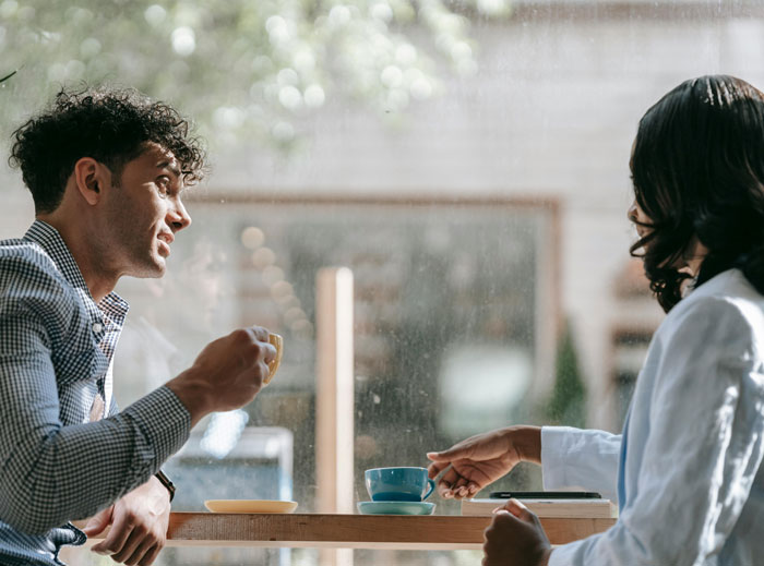Two people sitting by a window, engaged in conversation over coffee, possibly discussing a man's background.