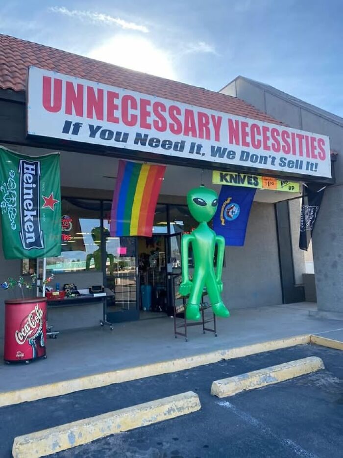 Storefront with a large green alien decoration and sign reading "Unnecessary Necessities," showcasing weird images.