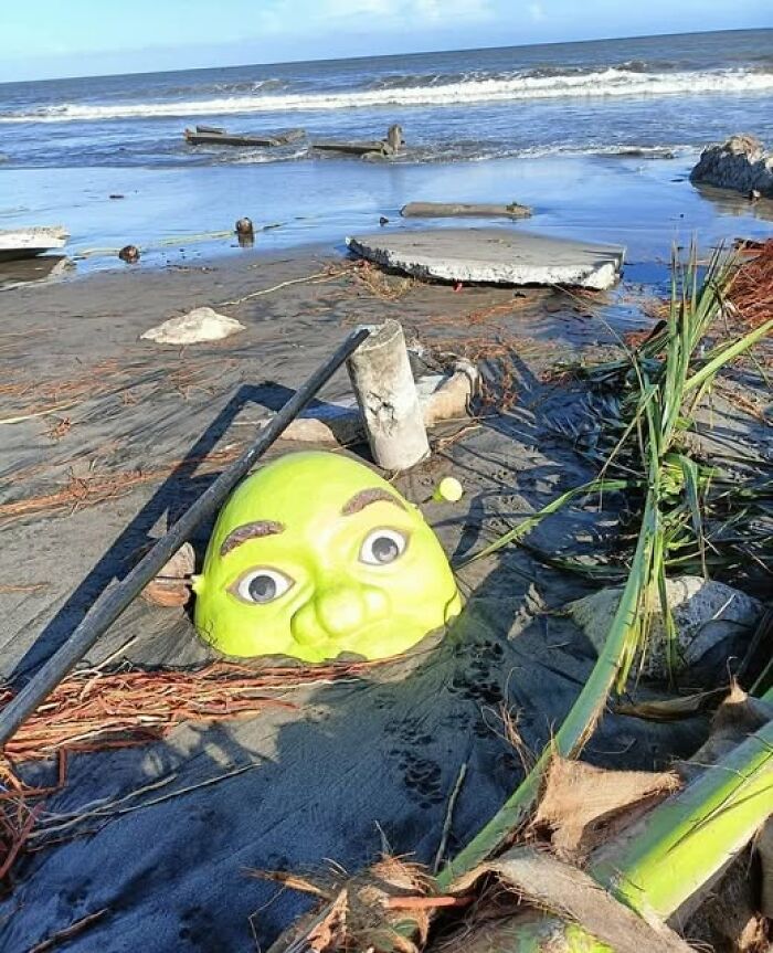 Weird image of a green mask partially buried in the sand on a beach with ocean waves in the background.