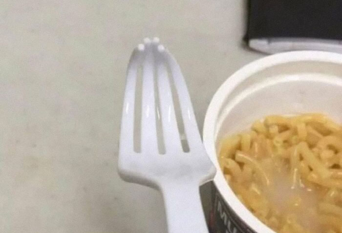 Weird image of a plastic fork missing prongs next to a cup of noodles.