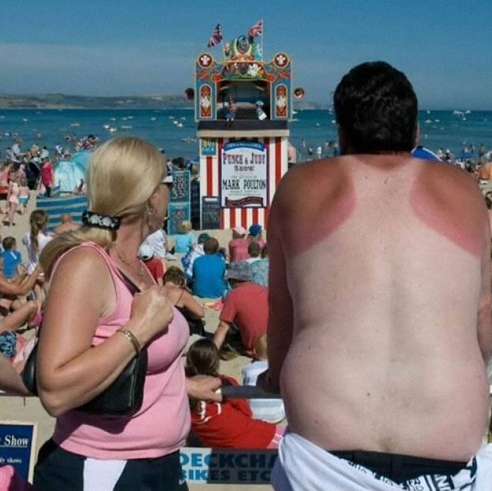 A sunburned man and woman watch a beach puppet show, creating a weird and amusing scene with a crowd in the background.