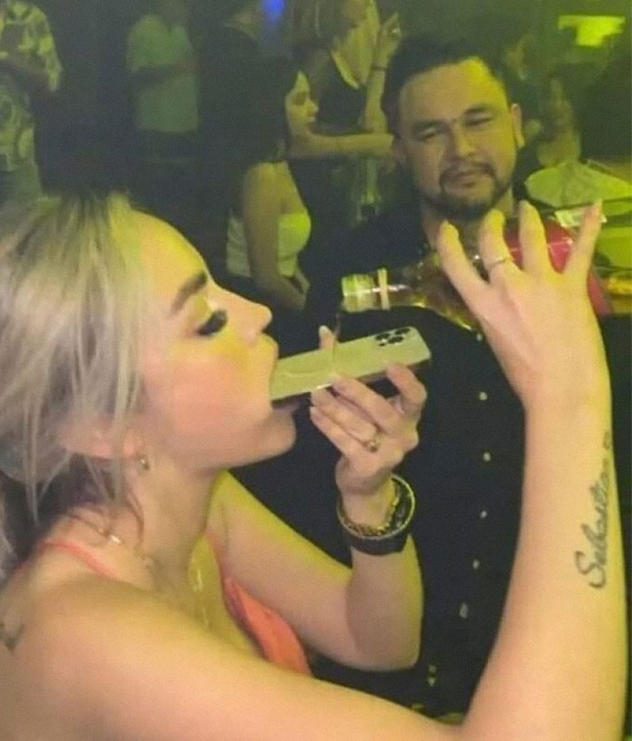 A woman using a phone as a funnel for a drink in a crowded club; an example of weird images.