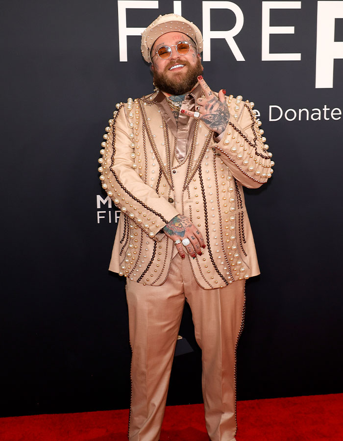 Man in pearl-studded beige suit, wearing sunglasses and hat, posing at 2025 Grammys red carpet.