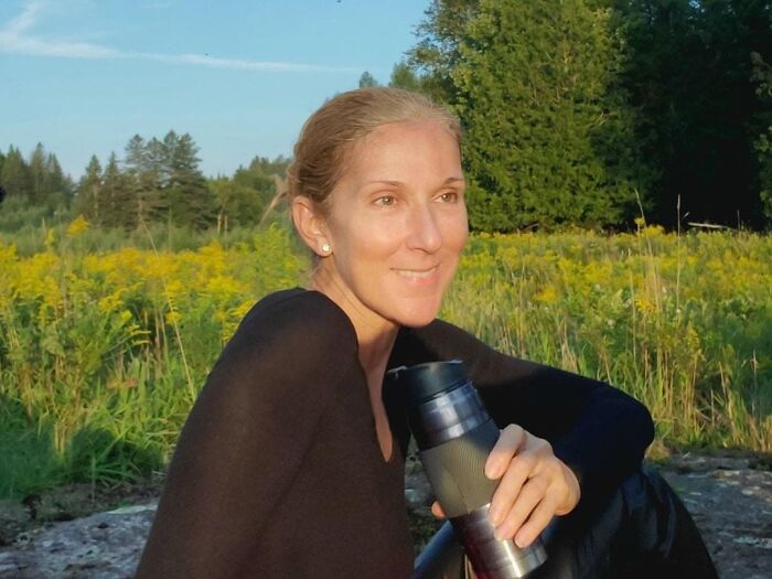 Celebrity without makeup, sitting outdoors in a field, holding a travel mug, wearing a black top, surrounded by greenery.