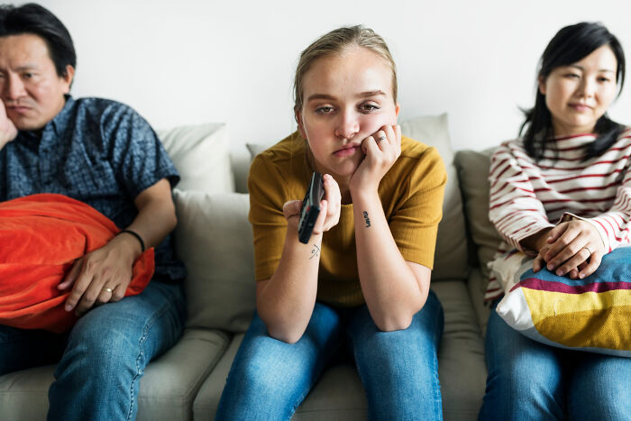 A young woman holding a remote looking bored sitting between two adults on a couch, relating to common experiences.