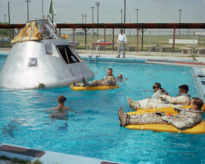 Astronauts in suits floating on rafts next to a space capsule in a pool, a rare historical training photo.