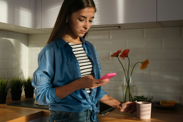 Woman in a blue shirt checking her phone in the kitchen, uncovering something in a man's background.
