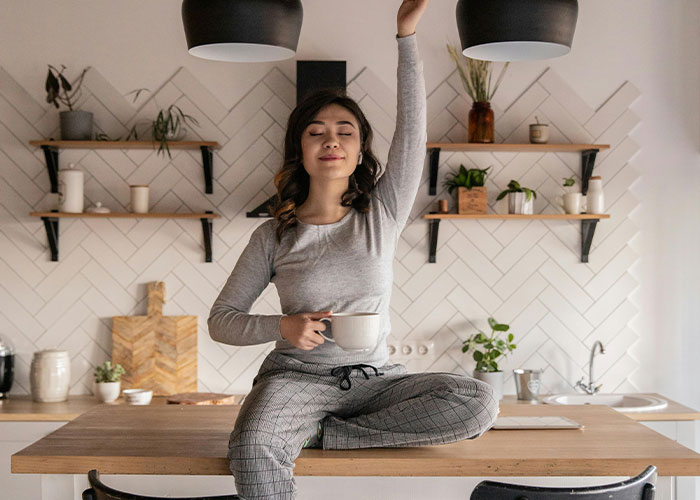 Woman enjoying a whimsical moment, sitting on a kitchen counter with a cup, surrounded by plants and cozy decor.