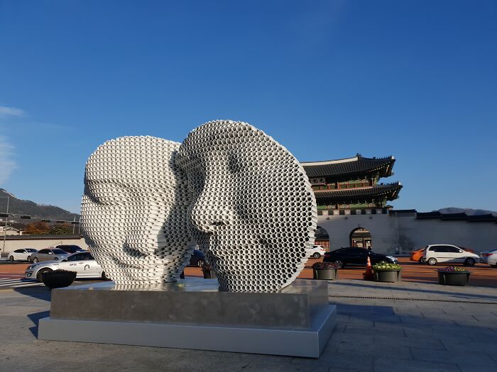 Metal pipe sculpture featuring two faces by Yi Chul Hee, displayed outdoors with a historical building in the background.
