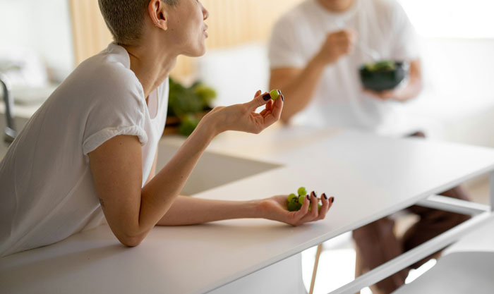Teen in a white shirt talking, holding a grape, seated at a table in a bright kitchen setting.