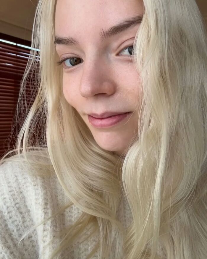 Celebrity without makeup, smiling with long blonde hair and wearing a cozy white sweater indoors.
