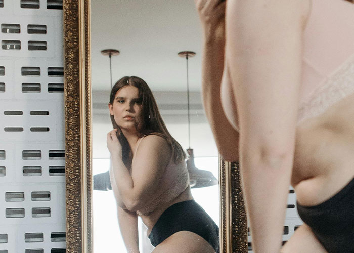 Woman in underwear thoughtfully looking at herself in a mirror, reflecting on challenges of being overweight.