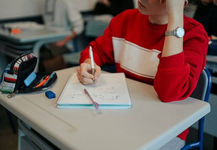 Student in a red sweater writing in a notebook during a class, with a focus on hilarious classroom moments.