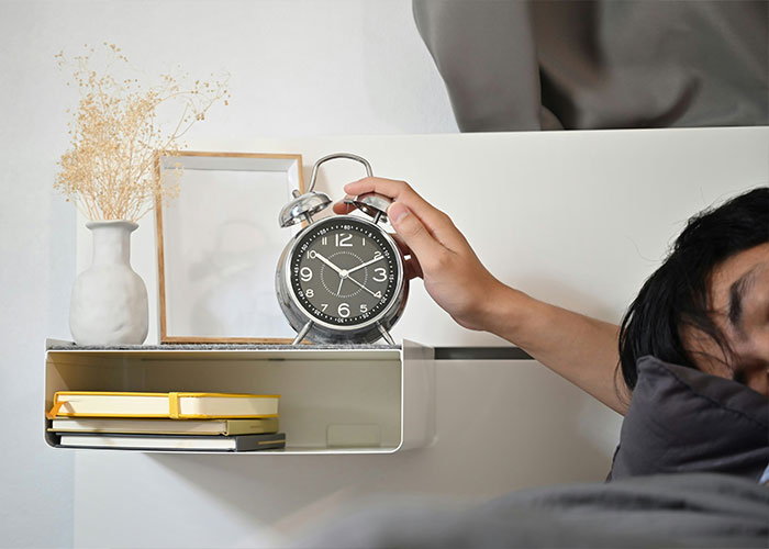 Person waking up and reaching for an alarm clock, with books and decor nearby, creating a whimsical morning scene.