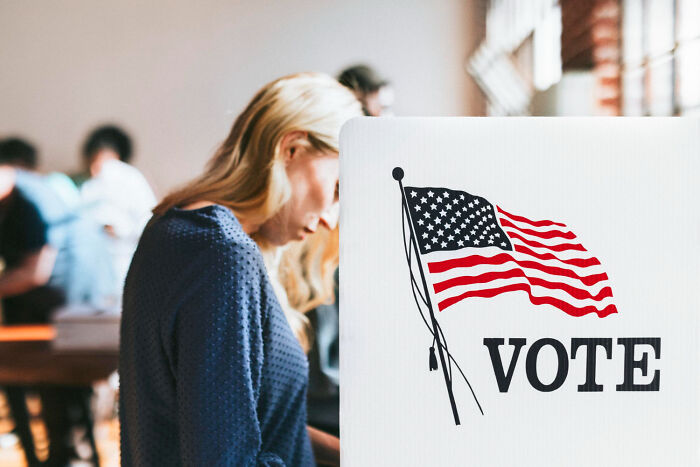 A woman voting in the USA, standing near a booth with the American flag and "VOTE" sign, highlighting a common practice.