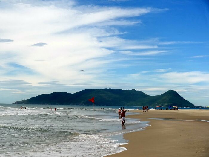 Retro Brazilian beach scene with people strolling along the sandy shore, mountains in the background, and a vibrant sky.