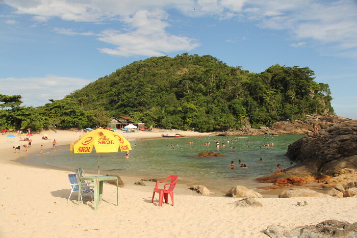 Retro Brazilian beach scene with a Skol umbrella, colorful chairs, and people swimming near a lush green hill.