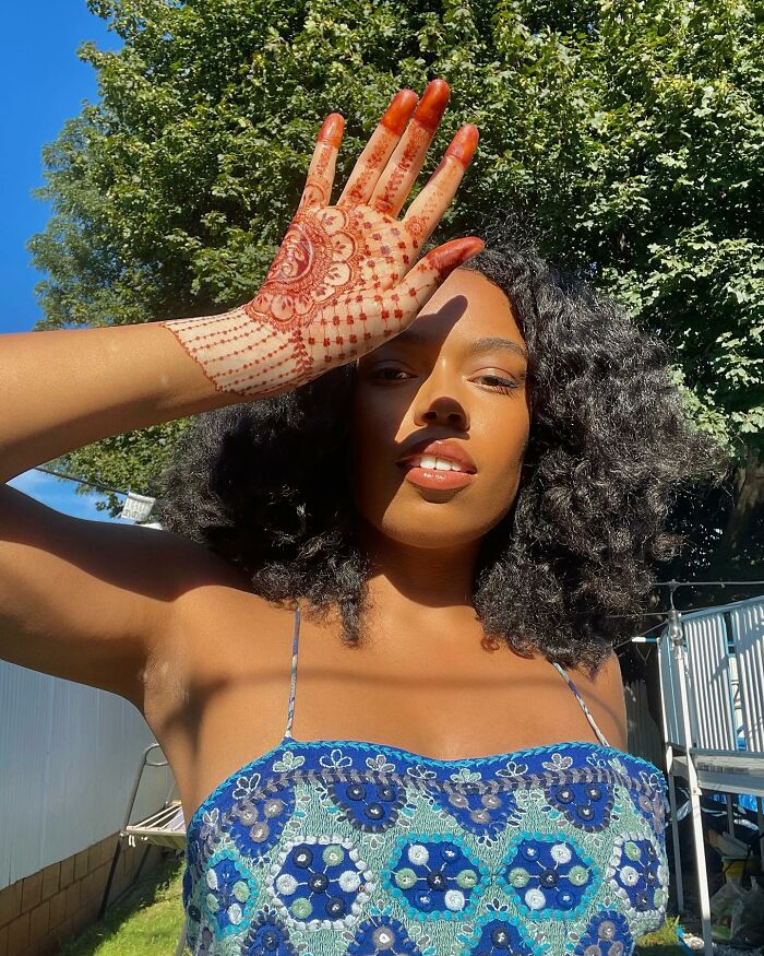 Woman outside with curly hair, wearing a blue top, showcasing beautiful body art on her hand in sunlight.