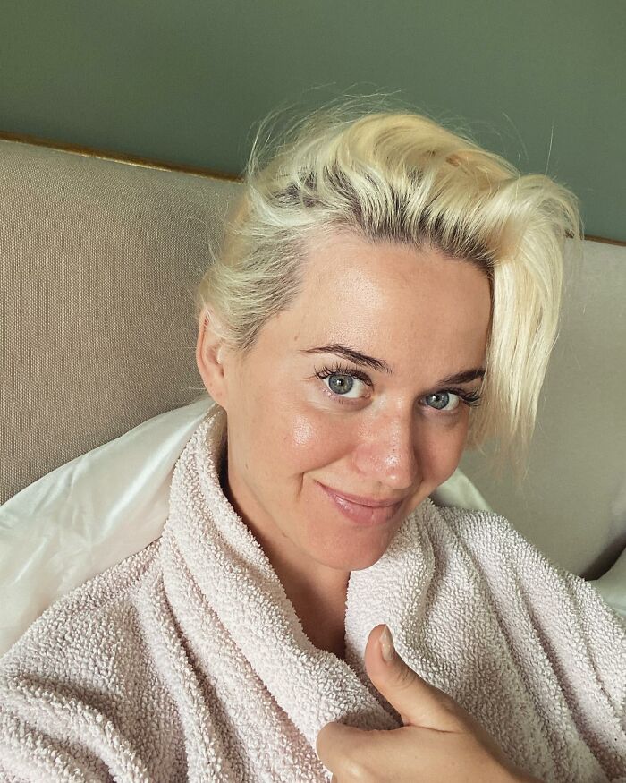 Blonde celebrity without makeup giving a thumbs-up, wearing a pink robe.