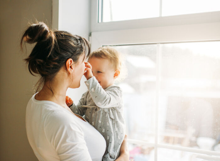 Mother holding baby near window in bright room, illustrating American norms of family bonding.