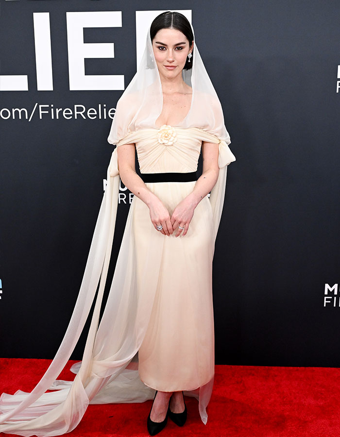 Elegant woman in a cream dress with veil on red carpet at the 2025 Grammys, accessorized with black heels and jewelry.