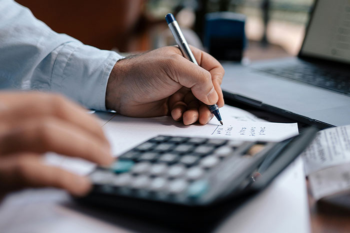 Person calculating expenses on paper with a pen, using a calculator and a laptop, related to safe side hustle ideas.