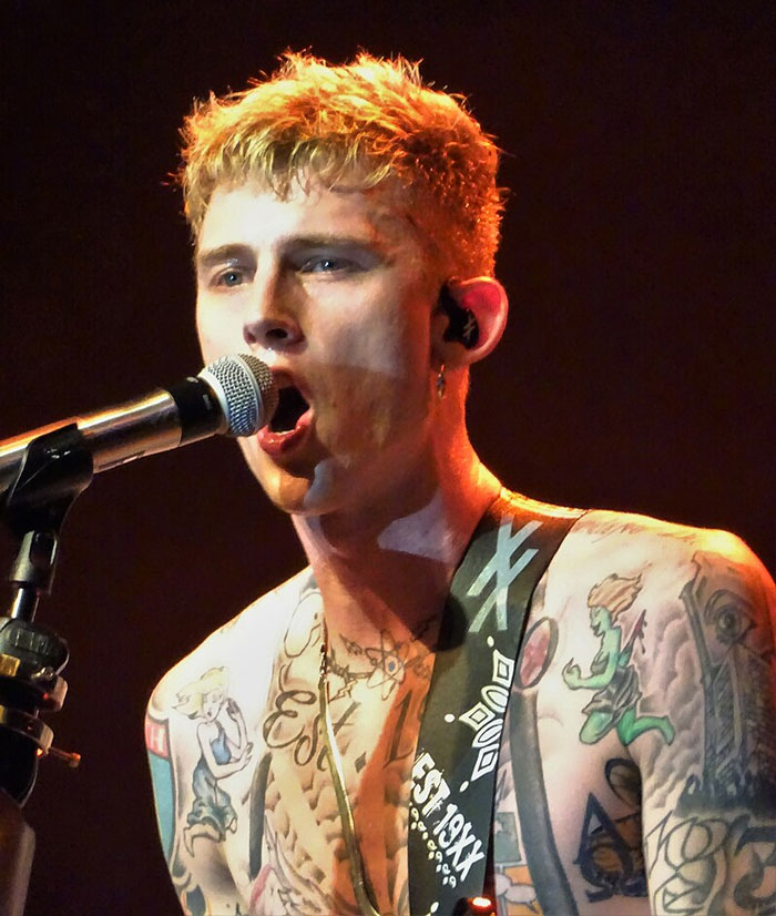 Singer performing live on stage, holding a microphone, with tattoos visible, framed as part of music discussion online.