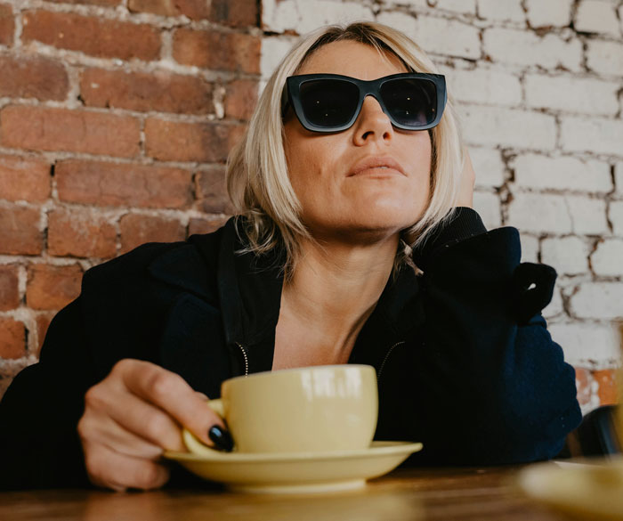 Rich person in sunglasses, holding a yellow cup, sitting against a brick wall, portraying a disconnect with reality.